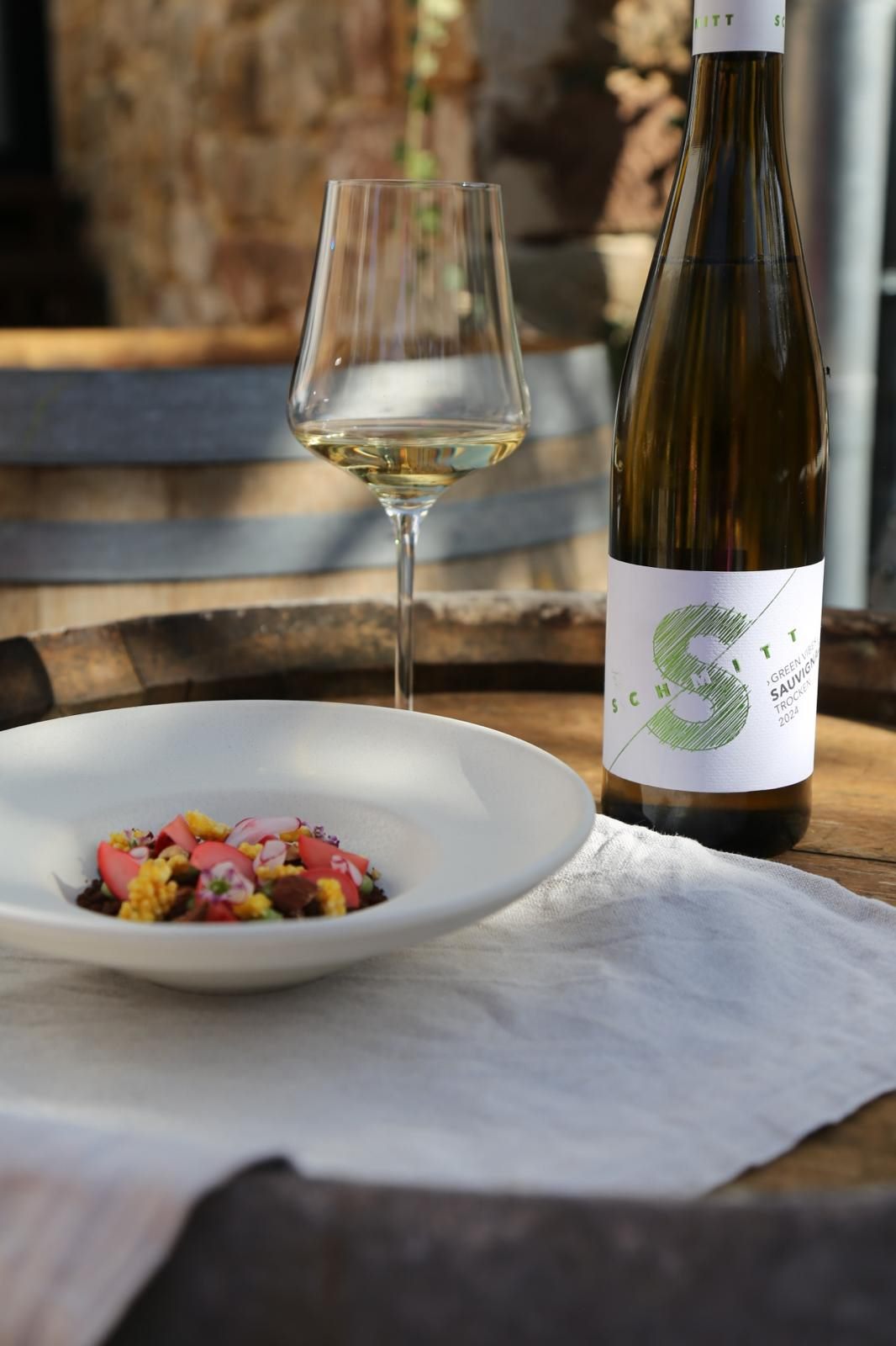Ein Gang auf dem Teller, daneben Glas und Flasche Sauvignon auf einem Weinfass